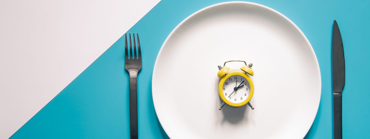 Alarm clock on a white plate with a knife and fork on blue background. Intermittent fasting, Ketogenic dieting, weight loss, meal plan, and healthy food concept.